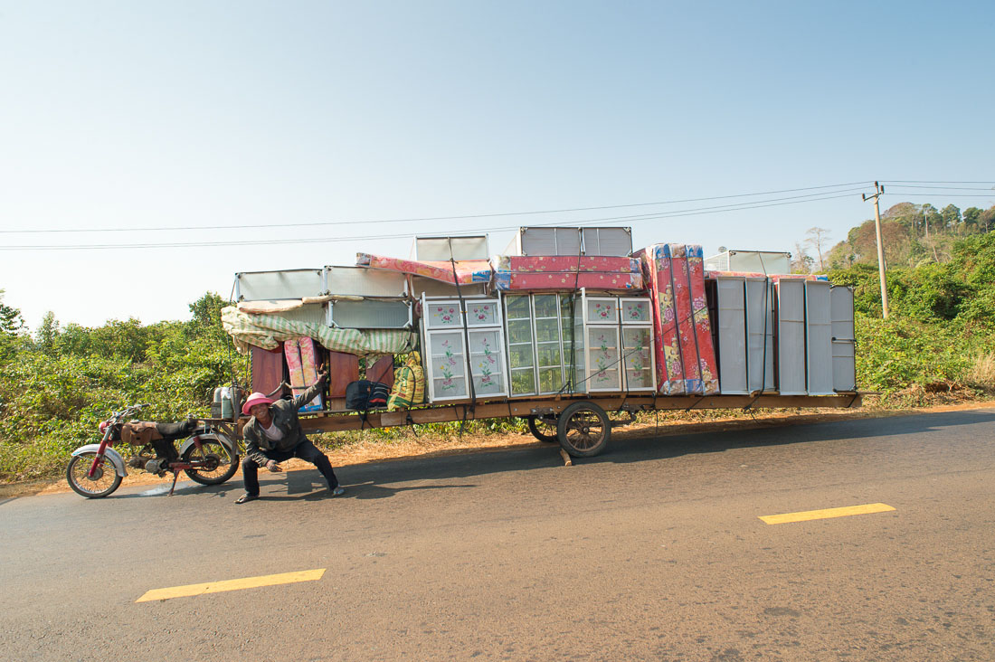 A really large and long trailer fully loaded with aluminum cabinet displays and mattresess, being towed by a small motorcycle. Kingdom of Cambodia, Indochina, South East Asia