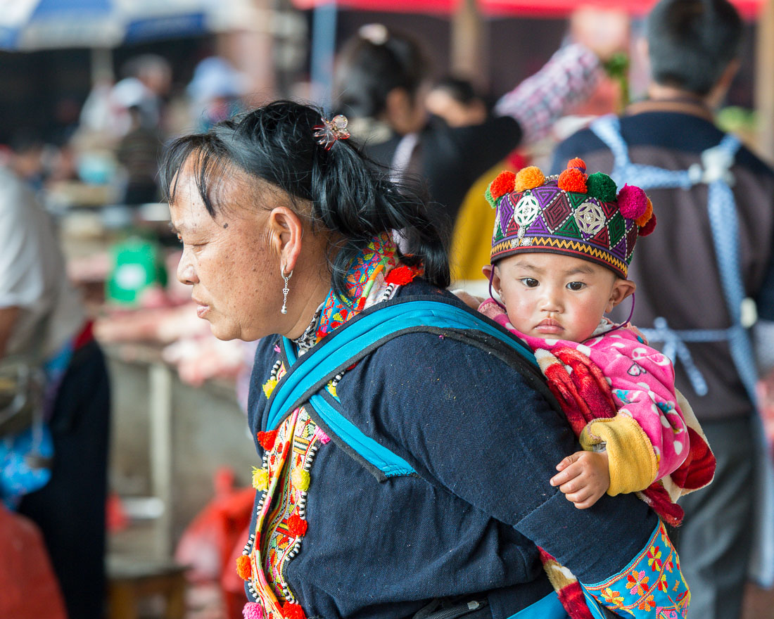 A grandma from the Yao ethnic minority people carrying her grandson, wearing traditional costume at the Jin Ping market, Yang Yang county, Yunnan Procince, China, Asia. Nikon D4, 70-200mm, f/2.8, VR II.