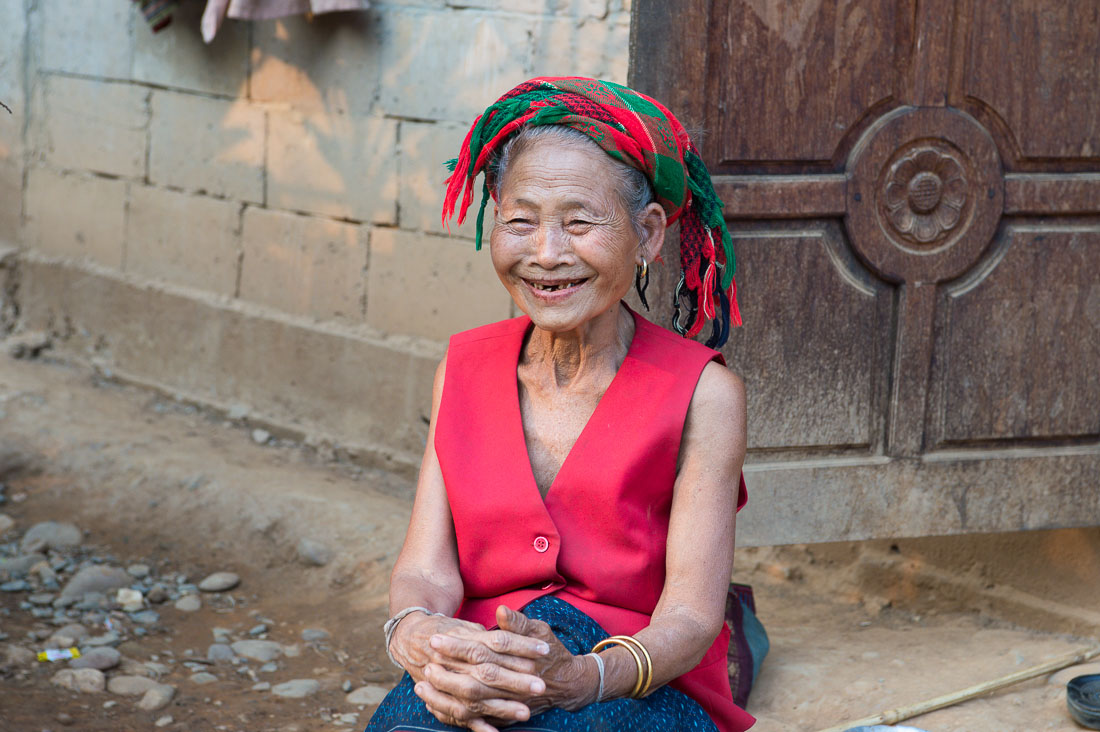 A very happy and smiling old woman from the Khamu people ethnic minority wearing her traditional costume, sitting in front oh her house; visible red stains on her teeth and the mouth area due to the chewing of paan (areca nuts and betel leaves). Lao PDR, Laos, Indochina, South East Asia