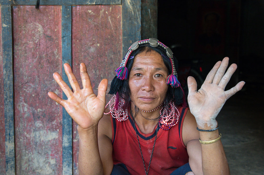 Woman from the Akha Korpean people ethnic minority wearing her traditional and elaborate hat sitting at her house door showing her hands dirthy from cement. Phicheu Mai village, Bokeo province, Lao PDR, Indochina, South East Asia.